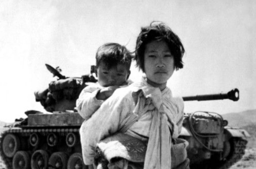 A child carrying a younger sibling in front of a tank, an archival photograph from the Korean War